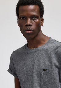 Nuori mustaihoinen mies, jolla on lyhyet kiharat hiukset ja harmaa pyöreäkauluksinen T-paita, jossa on pieni rintatasku, jossa lukee "Lee®".