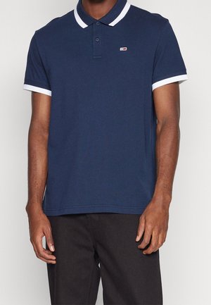 Polo shirt bleu marine avec des bordures blanches au col et aux manches, patte de boutonnage à trois boutons, et petit logo sur la poitrine ; fabriqué en coton doux.