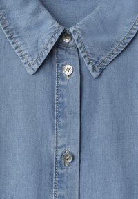 Chemise en denim bleu clair avec col pointu et trois boutons métalliques sur la patte de devant.