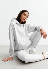 Jasnoszary hoodie z dużą kieszenią kangurką, połączony z dopasowanymi spodniami dresowymi. Model siedzi na gładkiej, neutralnej podłodze.