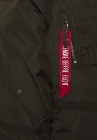 Tamno maslinasto zeleni bomber jaknu s rebrastim ovratnikom, džepom na patentni zatvarač i crvenom tkaninom oznakom s bijelim tekstom. Tekstuiran materijal.