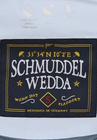 Marinblå tygetikett med vit och gul broderad text. Ikoner för vattentålig, vindresistent och syntetisk dunfunktion finns ovanför texten.