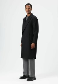 Zwarte wollen jas met een dubbelgeknoopt ontwerp, brede revers en zijzakken, gedragen over grijze broek en zwarte schoenen.