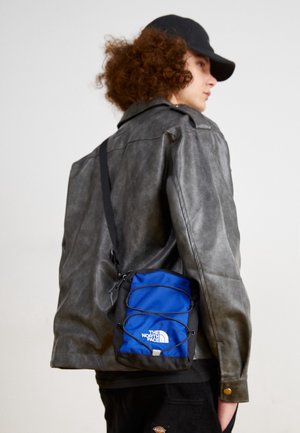 Person med krøllet hår iført en sort kasket og en grå læderjakke, der bærer en blå og sort The North Face skuldertaske.