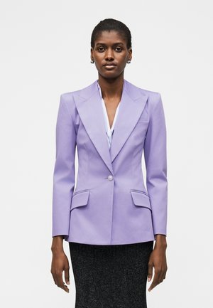 Naine, kes seisab näoga ette, kandes kohandatud lavendlivärvi bleiserit valge topi ja musta tekstuurse seeliku üle, seisab ühevärvilise tausta ees.