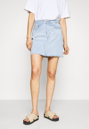 Personne portant une mini-jupe en jean bleu clair avec un ourlet brut, un t-shirt blanc et des sandales beige à enfiler sur un fond uni.