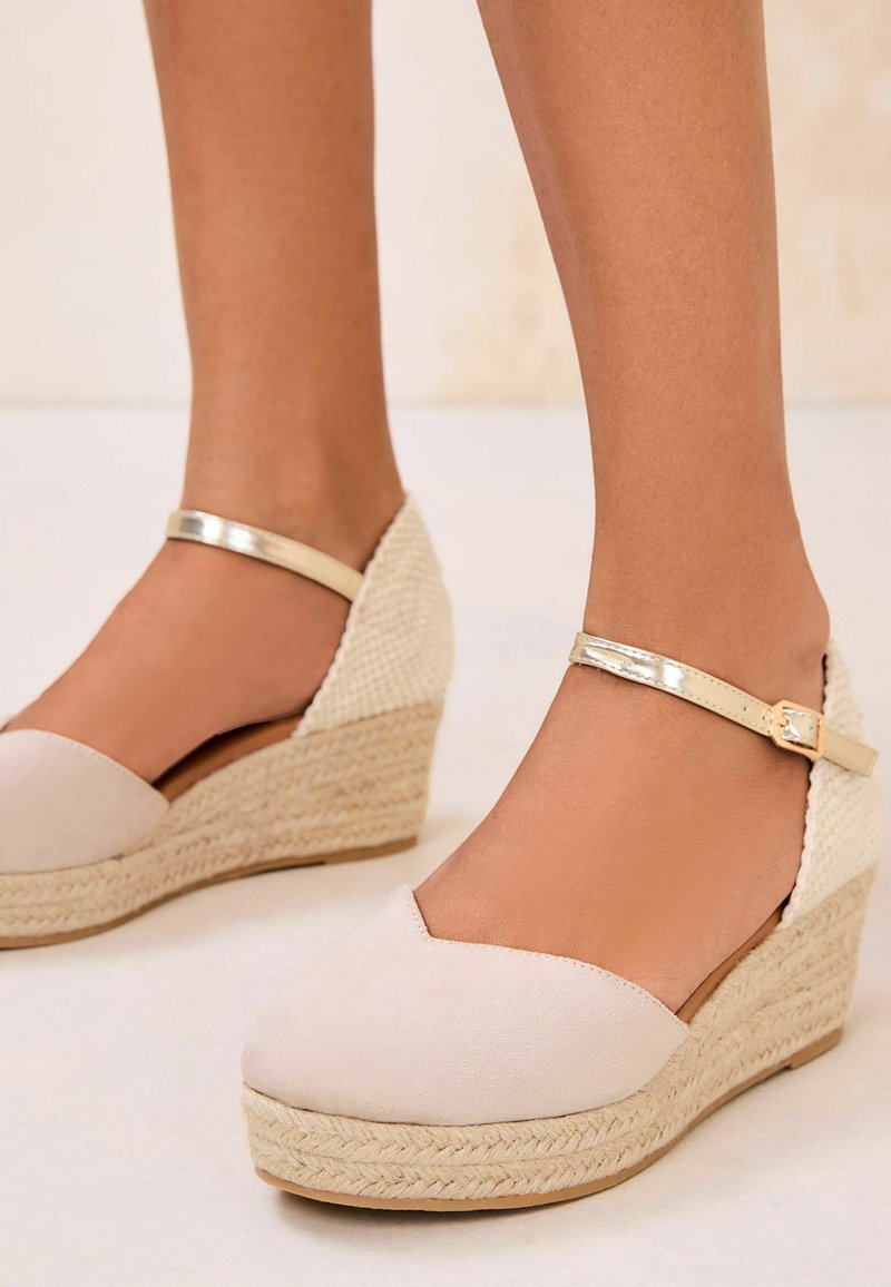 Beiget kiilakorkoiset espadrillot, joissa on punottu köysipohja ja metalliset nilkkaremmit, jalassa vaaleaa taustaa vasten.