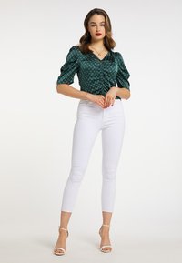 Grøn satinbluse med puffede ærmer og en rynket front, parret med tætsiddende hvide jeans og højhælede sandaler.