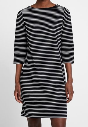 Mujer con vestido negro hasta la rodilla con rayas horizontales blancas y mangas tres cuartos, de pie frente a un fondo liso.