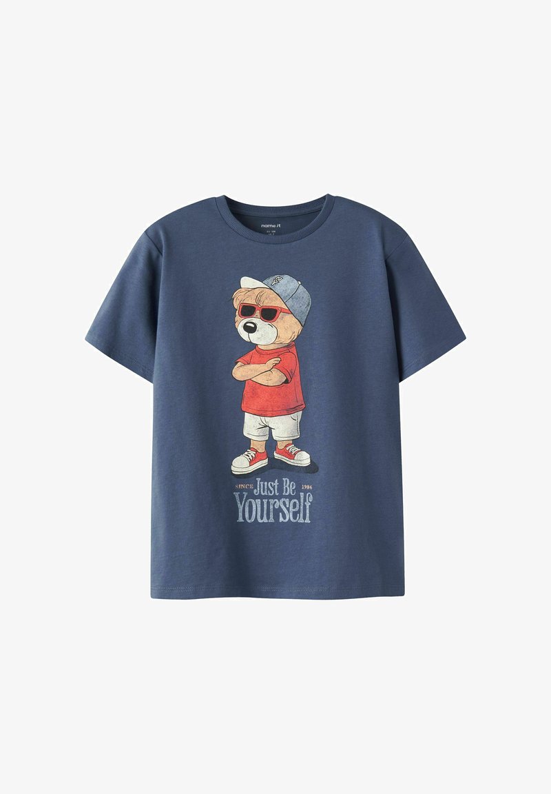 Μπλε σκούρο μπλουζάκι με γραφικό ενός αρκούδου με κόκκινα γυαλιά ηλίου, κόκκινο πουκάμισο, λευκές βερμούδες, αθλητικά παπούτσια και καπέλο, με κείμενο "Να είσαι απλώς ο εαυτός σου."