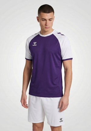 Man die een paarse en witte sportshirt met korte mouwen en witte korte broek draagt, staand tegen een effen lichte achtergrond, kijkend naar beneden.