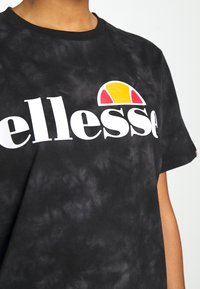 Camiseta negra con tie-dye, con un gran texto blanco "ellesse" y un logo amarillo y rojo en forma de media luna en el pecho, llevada por una persona con piel marrón clara.