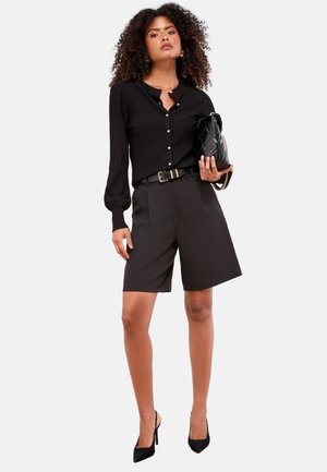 Femme aux cheveux bouclés portant un chemisier noir boutonné, un short ceinturé au genou, des talons noirs, et tenant un sac matelassé avec bandoulière chaîne.