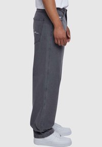 Pantaloni in denim grigi con vestibilità rilassata, caratterizzati da due tasche posteriori e un dettaglio del logo cucito, abbinati a sneakers bianche.