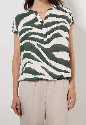 Kvinde iført kortærmet grøn og hvid zebrastribet bluse med knappet halsudskæring og lyse beige bukser mod en ensfarvet baggrund.