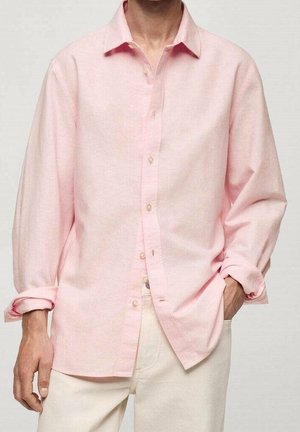 Mand iført en løs, lyserød skjorte med knapper og opkørte ærmer samt offwhite bukser, med den ene hånd i lommen mod en ensfarvet baggrund.