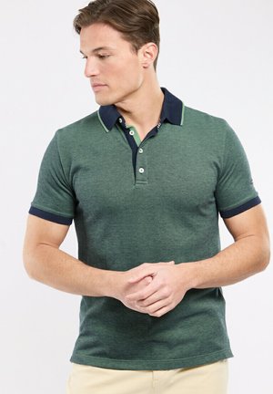 Homme portant un polo vert à manches courtes avec un col et des poignets de manches bleu marine, debout les mains jointes, regardant vers la gauche.