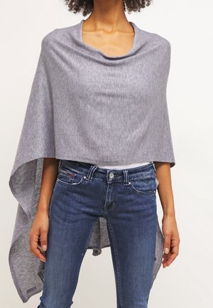 Frau trägt einen hellgrauen asymmetrischen Poncho über dunkelblauen Jeans, Hände entspannt seitlich an einem einfarbigen Hintergrund.