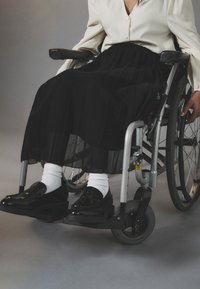 Jupe plissée noire, chemise blanche à boutonnage, chaussures noires et chaussettes blanches ; fauteuil roulant avec cadre argenté et roues noires.