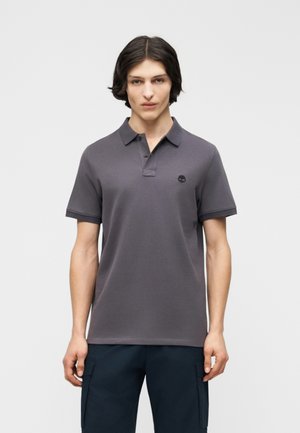 Junger Mann mit mittellangen dunklen Haaren, der ein dunkelgraues Poloshirt und schwarze Hosen trägt und vor einem schlichten hellen Hintergrund steht.