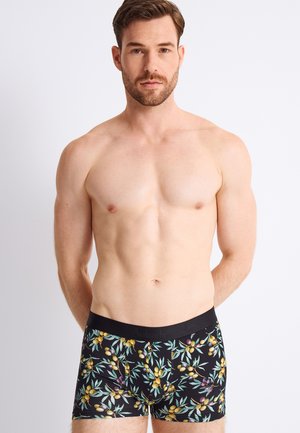 Homme torse nu aux cheveux courts et bruns, portant un boxer noir avec des motifs de branches d'olivier jaunes et violettes sur fond blanc.