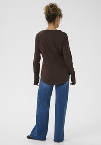 Langærmet brun skjorte med crew neck og afslappet pasform, sammen med brede blå jeans og sorte sko; set fra bagsiden.