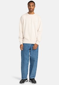 Sudadera beige de cuello redondo con puños y dobladillo acanalados, combinada con vaqueros de mezclilla azul holgados y zapatos deportivos negros.