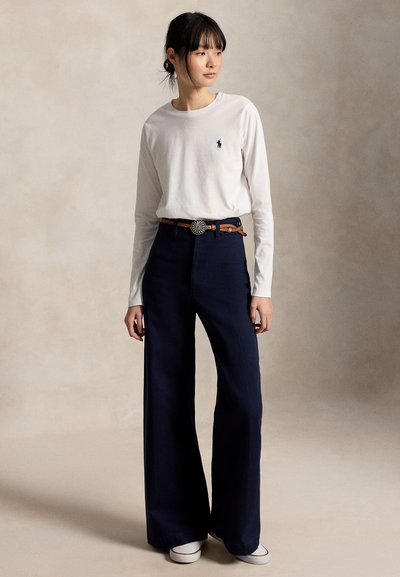 Γυναίκα όρθια, φοράει άσπρο μακρυμάνικο πουκάμισο με λογότυπο, σκούρο μπλε φαρδιά παντελόνια, καφέ ζώνη και άσπρα παπούτσια, κοιτάζει λίγο προς το πλάι.