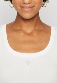 Hvid ribbet tanktop med en bred halsudskæring og en dekorativ trekant af sølvnitter på midten af brystet. Glat tekstur, tætsiddende design.
