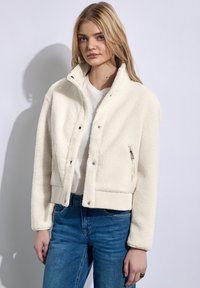Witte faux shearling jas met een cropped ontwerp, drukknoopjes, ritszak en geribbelde mouwen, gecombineerd met een effen wit t-shirt en blauwe jeans.
