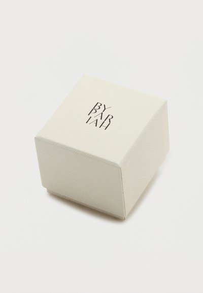 Malá béžová textúrovaná štvorcová krabička s čiernym nápisom značky „By Pariah“ na vrchu, umiestnená na bielom pozadí.
