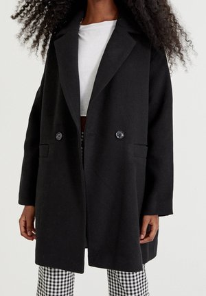 Femme portant un manteau noir à double boutonnage, un haut blanc et un pantalon à carreaux noir et blanc sur un fond clair uni.