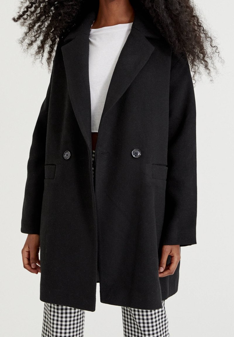Femme portant un manteau noir à double boutonnage, un haut blanc et un pantalon à carreaux noir et blanc sur un fond clair uni.