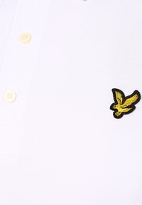 Polo alb cu un logo brodat al unui păun galben și negru, guler clasic și nasturi bej pe închidere.