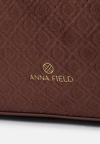 Flekkete brun stoff med et preget mønster og en gull "ANNA FIELD" logo. Designet er quilte for ekstra detaljer.