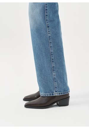 Jambes portant un jean droit bleu clair et des bottines en cuir marron foncé à talons blocs bas sur fond blanc.