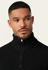 Schwarzer Strickpullover mit hohem Kragen und halbem Reißverschluss. Verfügt über eine gerippte Textur und subtile Nähdetails.