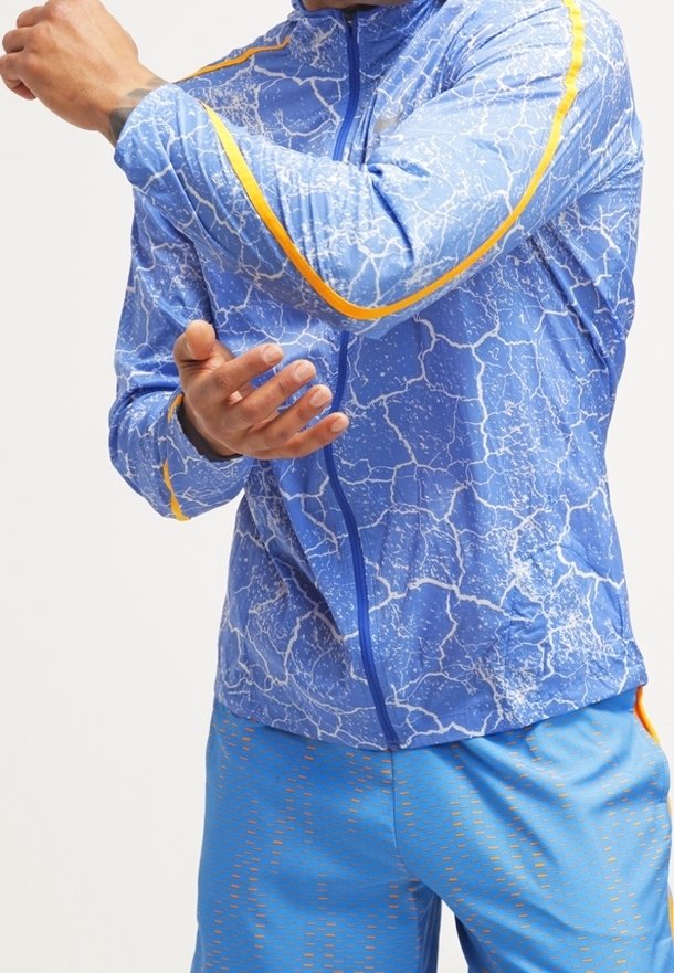 Veste de sport bleue avec un motif de texture fissurée, des accents jaunes et une fermeture éclair intégrale. Portée avec un short bleu assorti présentant des détails texturés.