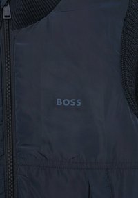 Gilet nero con una texture liscia, colletto a coste, chiusura con zip e un logo in rilievo nella zona del petto. Presenta un design a strati.