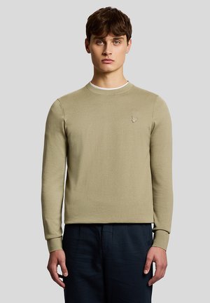Jeune homme portant un pull ras du cou beige clair avec un petit logo brodé sur la poitrine et un pantalon bleu marine foncé, debout devant un fond gris uni.