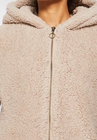 Beige fleece hættetrøje med en struktureret overflade, fuld lynlås og hætte. Har en gylden lynlåsdetalje og blødt, fluffy materiale.