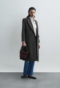 Dugi tamnosivi vuneni kaput s krojenjem na mjeru, dvostruko dugme sprijeda, uparen s prugastom košuljom, plavim trapericama i bordo torbom.