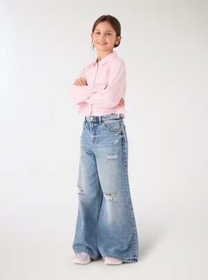 Ragazza giovane che sorride con le braccia incrociate, indossa una camicia rosa a righe, jeans larghi strappati e scarpe da ginnastica bianche su sfondo bianco.