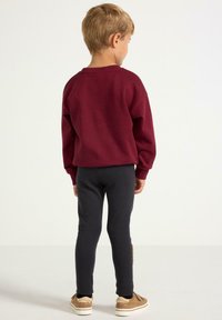 Punainen huppari raglan-hihoilla, yhdistetty tummanharmaisiin legginsseihin. Asusteena vaaleanruskeat kengät. Neutraali tausta.