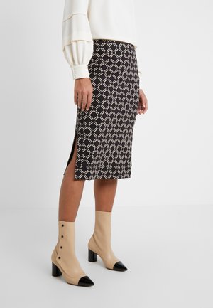 Kobieta ubrana w czarno-beżową midi spódnicę w kratę z bocznym rozcięciem, białą bluzkę oraz beżowe botki z czarnymi noskami i guzikami.