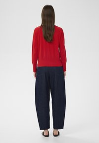 Pulover roșu tricotat cu mâneci lungi și tiv strâmt, asortat cu pantaloni loți din denim bleumarin, văzut din spate.