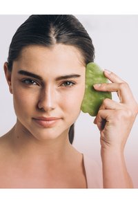 Grünes Jade-Gua Sha-Werkzeug, das an der Wange einer Frau gehalten wird, glatte Textur, natürliches Steinmaterial und konturierte Form sind sichtbar.