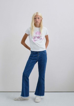 Camiseta blanca con gráfico de calavera rosa, combinada con vaqueros de mezclilla azules de corte acampanado con cierre de botones y dobladillo ligeramente deshilachado. Zapatillas blancas.