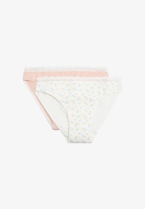 Mujer con cabello corto y castaño, vestida con un sujetador blanco y ropa interior rosa claro con encaje, de pie con los brazos levantados contra un fondo liso.