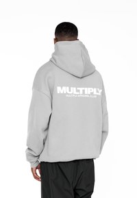 Felpa grigia oversize con un grande logo bianco "MULTIPLY" sulla schiena. Caratterizzata da un ampio cappuccio e maniche larghe. Abbinata a pantaloni neri.